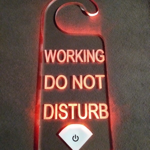 Puede incluir: Un colgador de puerta de acrílico rojo iluminado con el texto "WORKING DO NOT DISTURB" y un botón de encendido blanco.