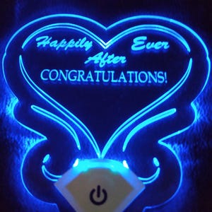 Könnte beinhalten: Ein blaues LED-Licht in Herzform mit dem Text "Happily After Ever Congratulations!" Das Licht hat einen rautenförmigen Ein-/Ausschalter.