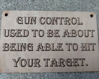 May include: A rectangular wooden sign with the engraved text "GUN CONTROL USED TO BE ABOUT BEING ABLE TO HIT YOUR TARGET." The sign has two pre-drilled holes at the top for hanging.