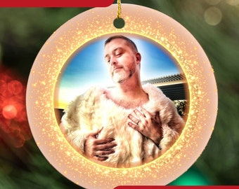 May include: A round, white Christmas ornament with a gold glitter border. The ornament features a photo of a person wearing a white furry coat. The text "Best Random Gift!" is at the top of the ornament and "Great for White Elephant Gift Exchange" is at the bottom.