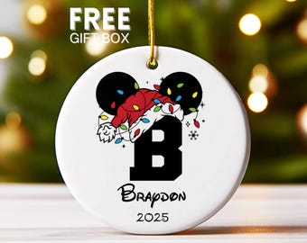 May include: White ceramic Christmas ornament with a black letter "B", a red Santa hat, and black mouse ears. The ornament has the name "Brapdon" and the year "2025" written on it. A gold string is attached for hanging.
