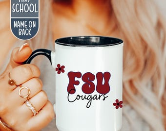 May include: White ceramic mug with a black rim and handle. The mug features the text "FSU Cougars" in maroon and black, with maroon flower accents. The mug is held by a person.