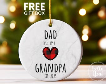 May include: A white, round Christmas ornament with the words "DAD EST. 1998" and "GRANDPA EST. 2025" in black. A red and black plaid heart is in the center. The ornament has a gold string for hanging.