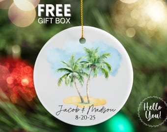 May include: White ceramic ornament featuring a watercolor illustration of two palm trees on a sandy beach under a blue sky. The ornament includes the names "Jacob & Madison" and the date "8-20-25". The text "FREE GIFT BOX" is at the top.