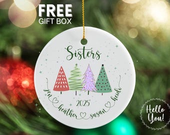 May include: A white ceramic Christmas ornament with a gold string. The ornament features the word "Sisters" above four colorful Christmas trees. Below the trees are the names Jen, Heather, Susan, and Heidi, with the year 2025.