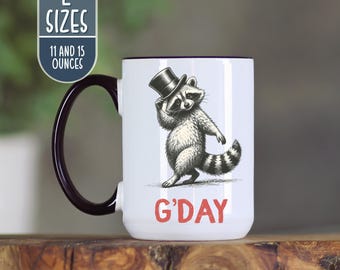 May include: White ceramic mug with a black handle and rim, featuring a raccoon wearing a top hat. The raccoon is holding its hat and the word "G'DAY" is printed in red. The mug is available in 11 and 15 ounce sizes.
