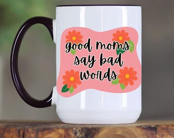 May include: White ceramic mug with a black handle and rim. The mug features a pink rectangle with orange flowers and the text "good moms say bad words" in black script. A fun gift idea.