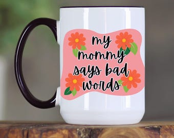 May include: White ceramic mug with a black handle and rim. The mug features a pink oval with orange flowers and the text "my mommy says bad words" in black script. The mug is on a wooden surface.