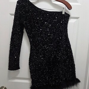 May include: A black one-shoulder mini dress covered in small, reflective sequins. The garment features a single long sleeve and a hemline trimmed with black feathers, creating a textured appearance throughout the entire piece.