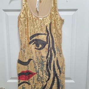 May include: A sleeveless tank dress covered in gold sequins featuring a large graphic design of a face in black sequins with red sequined lips. The dress has a scoop neckline and a beige lining, with a size XL tag visible at the collar.