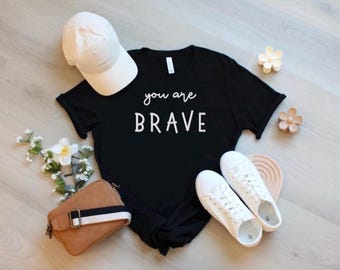 Op de afbeelding: Een zwart t-shirt met korte mouwen en de witte tekst you are BRAVE, een witte baseballpet, een paar witte sneakers, een bruine schoudertas met een zwart-witte gestreepte riem, twee kleine bloemvormige haarclips en een boeketje witte bloemen.