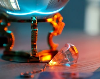 May include: A clear crystal pendulum with a silver chain hanging from a gold stand with a glass orb on top. The stand is on a wooden surface with a blue and orange glow.