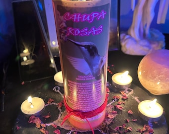 May include: A red and white candle with the words "CHUPA ROSAS" printed on the front. The candle is surrounded by seven small white candles and rose petals on a black slate surface. The candle has a red string tied around it and the text "VELADORA CHUPA ROSAS" is printed on the side.