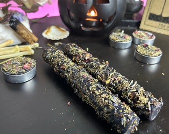 May include: Two black candles with dried herbs and flowers on a black surface. The candles are surrounded by small tea lights with dried herbs and flowers. A black jack-o'-lantern is in the background.