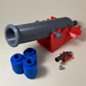 May include: A gray toy cannon with a red base, accompanied by three blue cylinders and a smaller black cannon. The toy cannon is a replica of a historical artillery piece. The cannon measures approximately 6 inches in length.