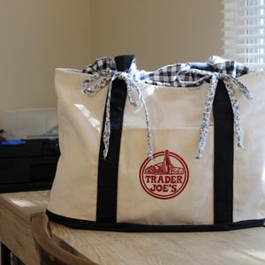 May include: Cream-colored tote bag with black trim and handles. The bag features a red Trader Joe's logo and is tied with blue and white floral ribbons. The top is lined with a blue and white checkered fabric.