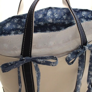 May include: Cream-colored tote bag with navy blue accents. The bag features navy blue handles and floral patterned ribbons tied into bows. The interior lining is a navy blue floral pattern. The bag has a white eyelet trim.