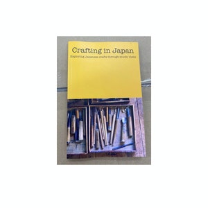 Op de afbeelding: Een boek getiteld "Crafting in Japan: Exploring Japanese crafts through studio visits" met een gele kaft en een foto van een houten kist gevuld met verschillende gereedschappen.