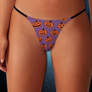 Puede incluir: Tanga morado con un patrón repetido de caras de calabaza naranja. La ropa interior tiene tirantes negros y está diseñada para Halloween. La imagen muestra la ropa interior en una persona.