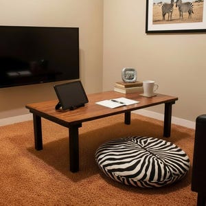 May include: A low, rectangular wooden table with black legs, a zebra-print floor cushion, and a framed picture of zebras. A black tablet sits on the table, along with a mug and books. A large black TV is mounted on the wall.