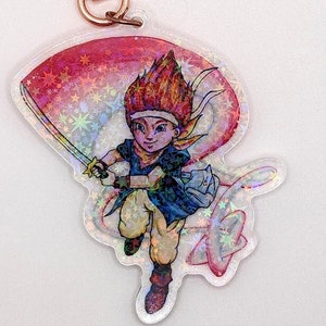 Puede incluir: Un llavero holográfico con un personaje de dibujos animados de pelo rojo, sosteniendo una espada y vistiendo una camisa azul y pantalones marrones. El personaje está rodeado de estrellas y un remolino rosa y rojo.