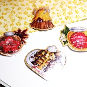 Op de afbeelding: Een verzameling stickers met voedselillustraties. Bevat een ahornsiroopfles, een pot honing, een stapel pannenkoeken met siroop en een grillig karakter. De stickers liggen op een wit oppervlak met een gele bloemenachtergrond.