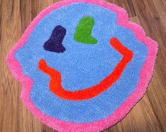 May include: A blue and pink smiley face rug with a bright orange smile and green and purple eyes.
