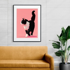 May include: A framed art print featuring a stylized black and white cat walking across a pink background. The cat has a white chest and paws, with a black tail and ears. The artwork is displayed on a white wall above a mustard-colored sofa.