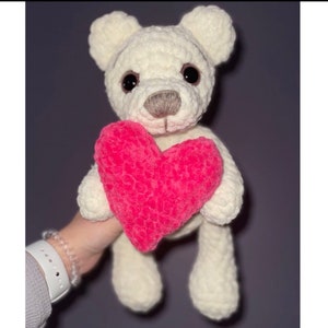 Könnte beinhalten: Ein weißer gehäkelter Teddybär, der ein rosafarbenes Plüschherz hält.