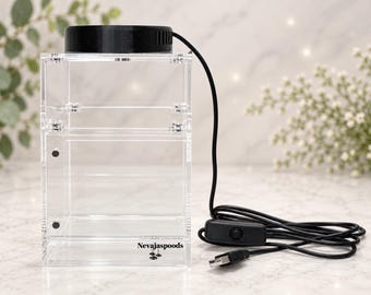 May include: A clear acrylic display box with a black circular top and a black USB power cord featuring an inline switch. The front of the box is printed with the text Nevajaspoods and a small spider icon. The structure is held together by silver screws.