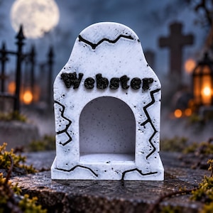 May include: A white, stone-textured tombstone-shaped decorative piece featuring a central arched opening and the name Webster in raised black lettering. The surface displays black, jagged crack patterns across the front face.