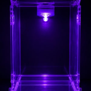 May include: A rectangular clear acrylic display case featuring a built-in circular light fixture at the top center. The unit emits a vibrant purple glow that illuminates the transparent structure, highlighting the edges and the internal space.