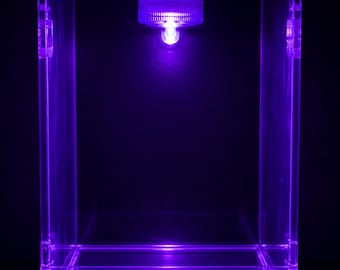 Può includere: Una teca rettangolare in acrilico trasparente dotata di un corpo illuminante circolare integrato nella parte superiore centrale. L'unità emette una vivace luce viola che illumina la struttura trasparente, evidenziando i bordi e lo spazio interno.