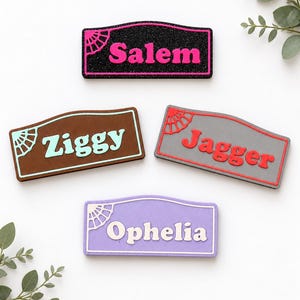 Puede incluir: Cuatro etiquetas rectangulares con bordes superiores arqueados, cada una con un diseño de abanico en una esquina. Las etiquetas muestran los nombres Salem en rosa sobre negro brillante, Ziggy en azul claro sobre marrón, Jagger en rojo sobre gris y Ophelia en blanco sobre morado.
