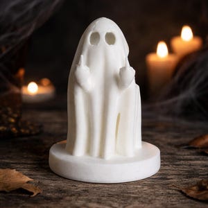 May include: A white ghost figurine standing on a circular base, featuring two hollow eye sockets and two arms raised with the middle fingers extended in a gesture. The object is rendered in a smooth, matte white material with a draped sheet appearance.