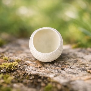 Op de afbeelding: Een kleine, bolvormige, gebroken witte kom met een getextureerd, mat buitenoppervlak en een gladde, holle binnenkant. Het object heeft een cirkelvormige opening aan de bovenkant en is gevormd als een uitgeholde schaal of open vat.