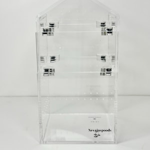 以下が含まれることがあります： 透明なアクリル製のジュエリーオーガナイザー。家型のトップが付いています。オーガナイザーには複数の棚とヒンジがあります。「Nevajaspoods」の文字が前面に印刷されています。イヤリング、ネックレス、その他のジュエリーを収納するように設計されています。