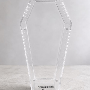 May include: A clear acrylic display case shaped like a coffin with a hollow center and a series of small holes along the inner edges. The base features the black text Nevajaspoods above a small graphic of two hanging eyes.