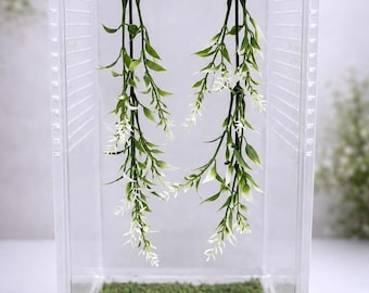 May include: A clear acrylic enclosure featuring a rectangular structure with ventilation holes on the sides. The interior floor is covered in green moss, and two artificial green and white hanging plants are suspended from the top ceiling.