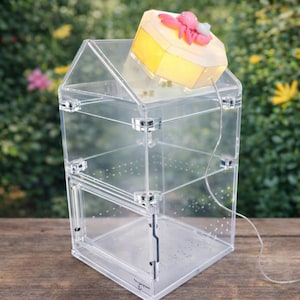 May include: A clear acrylic terrarium with a house-like shape, featuring a hinged door and multiple levels. A decorative, illuminated box with pink and white accents sits on top. A wire extends from the box.