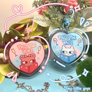 Peut inclure: Deux porte-clés en acrylique en forme de cœur, de couleur rose et bleue. Le porte-clés rose présente un ours brun de dessin animé portant un nœud rose et lisant un livre avec le texte "Pass the Yuri". Le porte-clés bleu présente un chat blanc de dessin animé portant un bandeau bleu et lisant un livre avec le texte "Pass the Yaoi".