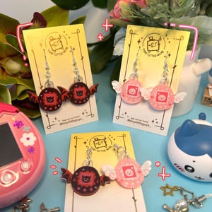 Puede incluir: Tres pares de pendientes originales en tarjetas amarillas. Dos pares presentan un diseño circular con alas de murciélago, uno en rojo y otro en rosa con alas blancas. El tercer par tiene un diseño circular rosa con alas blancas. El texto "tiny-little-guys" es visible.