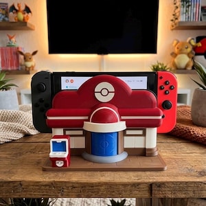 May include: A red, white, and blue gaming console dock shaped like a building with a circular emblem on the front. The dock holds a black and red handheld gaming console with a screen visible. A small red and white computer terminal sits on the base.