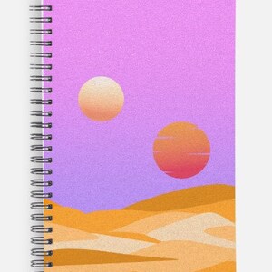 May include: Spiral-bound notebook featuring a desert landscape design. The cover art depicts two suns in a purple sky above sand dunes. The notebook has a silver spiral binding.