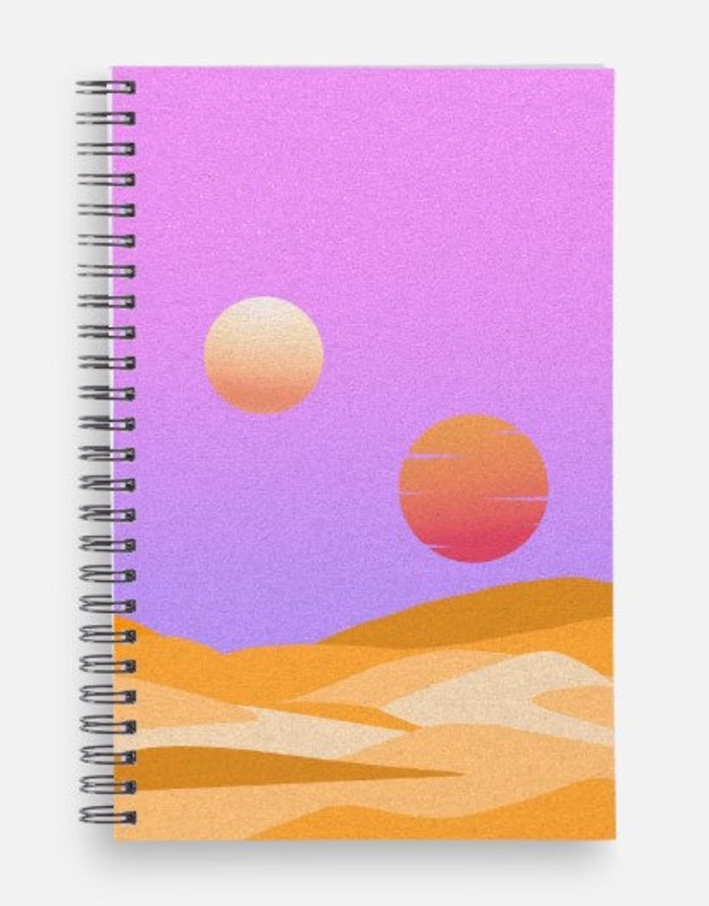 May include: Spiral-bound notebook featuring a desert landscape design. The cover art depicts two suns in a purple sky above sand dunes. The notebook has a silver spiral binding.