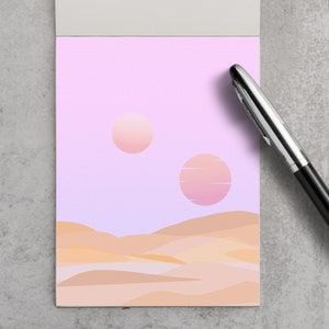 May include: A notepad with a landscape illustration featuring two suns in a pink and purple sky above tan-colored dunes. A black and silver pen rests on the right side of the notepad.