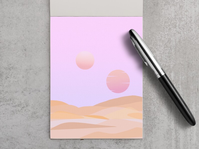 May include: A notepad with a landscape illustration featuring two suns in a pink and purple sky above tan-colored dunes. A black and silver pen rests on the right side of the notepad.