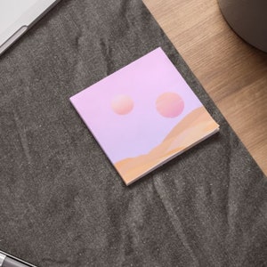 May include: A square notepad with a pastel landscape design. The design features two peach-colored orbs in a lavender sky above tan-colored dunes. The notepad is on a dark gray surface, near a laptop and pen.