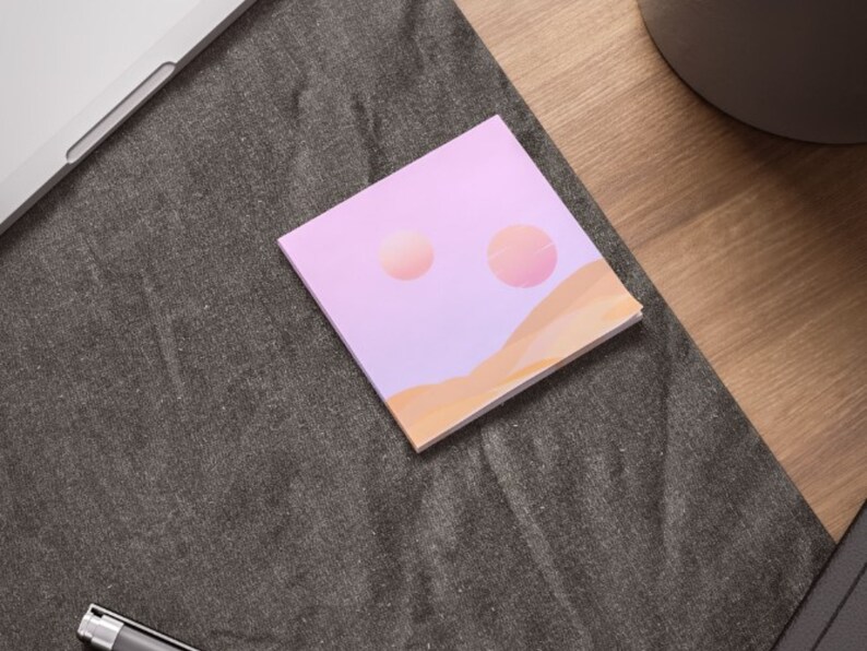 May include: A square notepad with a pastel landscape design. The design features two peach-colored orbs in a lavender sky above tan-colored dunes. The notepad is on a dark gray surface, near a laptop and pen.