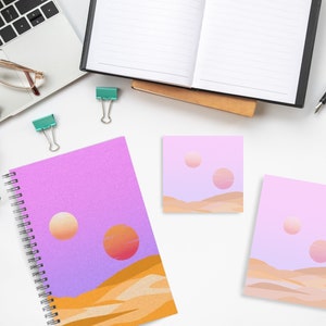 May include: A spiral-bound notebook, sticky notes, and a card featuring a desert landscape with two suns in shades of orange and pink. The notebook and stationery have a purple and orange color scheme. Other office supplies are also visible.
